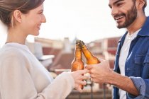 Jovens amigos encantados em roupas casuais que cercam garrafas de cerveja enquanto esfriam no terraço juntos — Fotografia de Stock