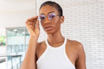 Впевнена афро-американська жінка з коротким волоссям в стильному одязі з модними окулярами стоять на вулиці біля білої цегляної стіни. — стокове фото