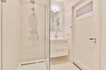 Moderno cuarto de baño interior con pared de vidrio contra lavabo debajo del espejo y lámpara brillante en casa - foto de stock
