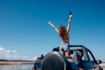 Back view of excited woman in summer outfit raising hand of beer while standing on roof of safari car on shore of river — Stock Photo