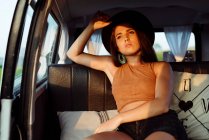Atractiva chica morena con sombrero dentro de una furgoneta vintage y tumbada en el asiento en un día soleado - foto de stock