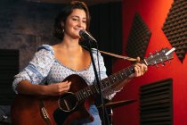 Уверенная леди с гитарой поет в микрофон, исполняя песню в ярком клубе — стоковое фото