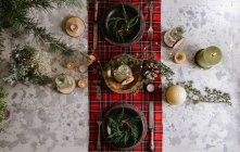 Вид на сервировку рождественского стола с венком на тарелке, декоративные деревянные украшения и красную клетчатую скатерть с желтыми огнями на заднем плане — стоковое фото
