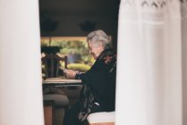 Вид збоку усміхненої літньої жінки в теплому одязі, що сидить за столом з таблеткою і чашкою чаю, дивлячись на екран — стокове фото