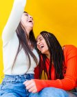 Счастливые подруги в разноцветных свитерах и джинсах сидят и смеются на желтом фоне — стоковое фото