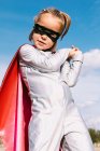 Знизу мила дитина в маскарадному костюмі супергероя і масці для очей, що стоїть на тлі блакитного неба і дивиться на камеру — стокове фото