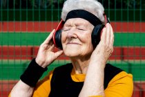 Взрослая женщина с проколотым носом в активной одежде слушает песни в наушниках, стоя на спортивной площадке возле сетки — стоковое фото