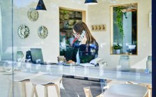 Durch das Glas einer Geschäftsfrau, die am Tisch mit Kaffee und Croissant neben dem Laptop auf dem Smartphone spricht, während sie aus der Ferne arbeitet — Stockfoto