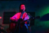 Confident lady with guitar with eyes closed singing in mic while performing song in bright club with neon light — Stock Photo