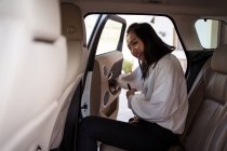 Позитивний етнічний пасажир в офіційному вбранні зі смартфоном, що входить в машину на задньому сидінні — стокове фото