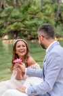 Couple marié positif en tenue de mariage et lunettes de soleil assis sur un escalier en pierre près du lac et des palmiers verts et des plantes tout en se regardant et en donnant des fleurs — Photo de stock