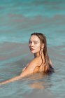 Mujer desnuda con el pelo mojado de pie en agua de mar mientras mira a la cámara sobre el hombro en la luz del día - foto de stock