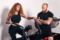 Deportista concentrada con el pelo largo rizado sentado en la máquina de ciclismo y haciendo ejercicio cardiovascular con entrenador personal en el gimnasio - foto de stock
