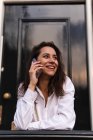 Seitenansicht einer glücklichen jungen Frau in lässiger Kleidung, die in der Nähe des Eingangs des Gebäudes steht und beim Telefonieren am Geländer lehnt — Stockfoto