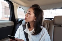 Asian female passenger in formal clothes browsing cellphone while sitting on backseat in taxi and looking away with interest — Stock Photo