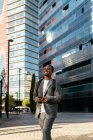 Улыбающийся афроамериканский бизнесмен в формальной одежде с рюкзаком просматривает мобильный телефон в центре города и смотрит прочь с зубастой улыбкой — стоковое фото