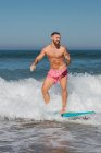 Активний чоловік у шортах для плавання стоїть на дошці для серфінгу під час махаючого моря в тропічному курорті в сонячний літній день — стокове фото