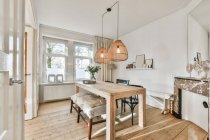 Design criativo da sala de jantar com banco e mesa de madeira sob lâmpadas penduradas contra janela na casa — Fotografia de Stock