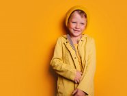Happy boy in vivid yellow raincoat jacket and beanie hat laughing and looking away against yellow background in studio — Stock Photo