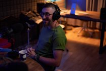 Joven positivo barbudo milenial masculino en gafas y auriculares sonriendo y hablando en micrófono mientras graba podcast en estudio oscuro - foto de stock