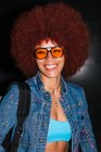 Donna sorridente con acconciatura afro e vestito alla moda e occhiali da sole che guarda la fotocamera mentre in piedi su sfondo nero con zaino in serata — Foto stock