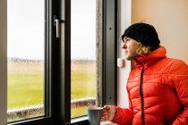 Calma joven viajero masculino en chaqueta roja caliente y sombrero beber taza de café caliente y mirando por la ventana de pie en la cocina en casa en el día lluvioso en el campo - foto de stock