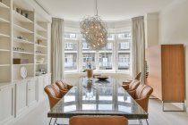 Table longue et chaises en cuir situées près des étagères avec décorations et fenêtre avec rideaux dans la salle à manger spacieuse — Photo de stock