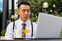 Joven empresario masculino étnico serio con tarjeta de crédito sentado en la mesa de la cafetería urbana con netbook y taza de café mientras hace compras en línea - foto de stock