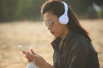Side view of serious Asian female in sunglasses text messaging on cellphone while listening to music in headphones on sandy shore — Stock Photo