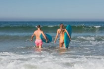 Vista trasera de cuerpo completo de una pareja deportiva en traje de baño con tablas de surf preparándose para surfear en el mar ondulado durante el entrenamiento ion ocean - foto de stock