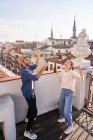 Full body of cheerful friends raising bottles of beer while laughing and dancing on balcony in old city — Stock Photo
