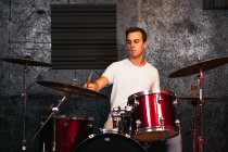 Concentrated young male musician in casual clothes playing crash cymbal in bright club near gray wall — Stock Photo
