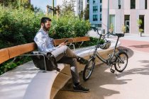Visão lateral do corpo inteiro do gerente masculino sentado no banco perto da bicicleta e lendo mensagens no smartphone no parque — Fotografia de Stock