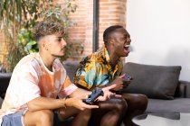 Amigos multirraciais alegres com gamepads em mãos sentadas no sofá enquanto jogam videogame juntos na sala de estar leve com planta verde — Fotografia de Stock