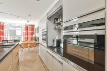 Cozinha moderna com bancadas de pedra preta e construído em aparelhos brilhantes em espaçoso apartamento leve com cadeiras à mesa na sala de jantar com janela — Fotografia de Stock
