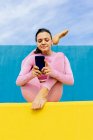 Положительная тонкая женщина сообщения на мобильный телефон, сидя в вариации сидячей колыбели позы во время занятий йогой Hindolasana на блю и желтый фон — стоковое фото