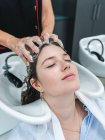 Cultive o mestre irreconhecível que lava o cabelo escuro do cliente feminino com olhos fechados no salão de beleza — Fotografia de Stock