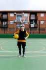 Mulher madura positiva em activewear e headband olhando para a câmera enquanto está de pé com bola durante o jogo de basquete — Fotografia de Stock