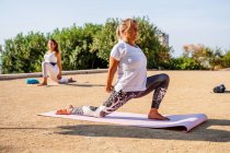 Side view of calm woman in activewear performing crescent lunge on knee with hands interlaced on mat during yoga practice with instructor in park in sunny day — Stock Photo