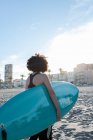 Vista posteriore di surfista donna irriconoscibile in muta con tavola da surf in piedi guardando lontano sulla riva del mare lavata dal mare ondulato — Foto stock