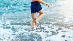 Barefoot cropped unrecognizable female traveler running along sandy beach washed by foamy waves in windy weather — Stock Photo