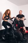 Deportista concentrada con el pelo largo rizado sentado en la máquina de ciclismo y haciendo ejercicio cardiovascular con entrenador personal en el gimnasio - foto de stock