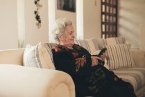 Mujer mayor enfocada con cabello gris descansando en el sofá y leyendo e libro en la tableta en la sala de estar en casa - foto de stock