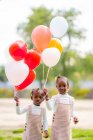 Счастливые афроамериканские младшие сестры в подобных платьях стоят с красочными воздушными шарами в руках на зеленой траве в парке при дневном свете — стоковое фото