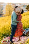 Side view of couple kissing and embracing while covering face with hat in blooming meadow in sunny day — Stock Photo