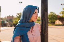 Vista lateral del pañuelo tradicional femenino musulmán positivo y mirando hacia otro lado en el día soleado en la ciudad - foto de stock