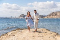 Повне тіло веселої весільної пари босоніж, що біжить на березі моря, насолоджуючись весільним днем на сонячній природі — стокове фото