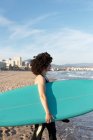 Vista lateral da jovem surfista em fato de mergulho com prancha de surf em pé olhando para longe na praia lavada pelo mar ondulado — Fotografia de Stock