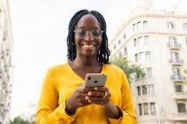 Positive African American female in eyeglasses text messaging on cellphone while standing on street with residential buildings on street in city — Stock Photo