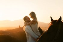 Vista lateral da fêmea feliz admirando o pôr do sol sobre as montanhas ao ser criado pelo homem amoroso entre cavalos calmos no campo da Turquia — Fotografia de Stock